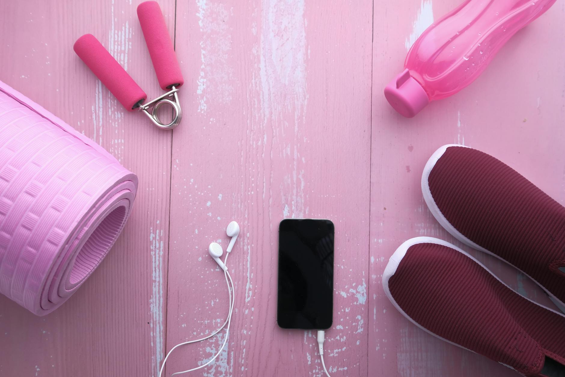 Akcesoria fitness na różowym tle: mata, buty, bidon i smartfon